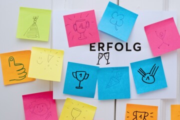 Bunte Notizzettel mit Erfolgssymbolen umgeben das Wort "ERFOLG" auf weißem Papier.